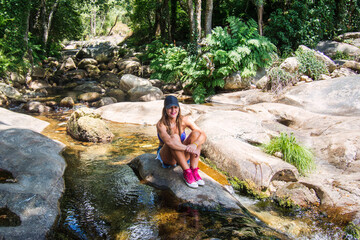 Woman Sitting On A Rock Surrounded By A Spectacular Natural Landscape. Young Girl Who Is In Contact With Nature, She Is Next To A River That Has Water Currents.