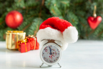 Christmas and New Year composition with a clock in a Santa hat showing the eve of the holiday, Fir tree branches, Holiday decorations and gifts