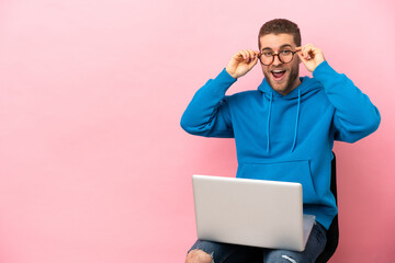 Young man sitting on a chair with laptop with glasses and surprised