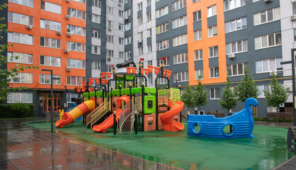 A courtyard of high-rise buildings with a new modern colorful and large playground on a rainy summer day without people. Empty outdoor playground. A place for children's games and sports.