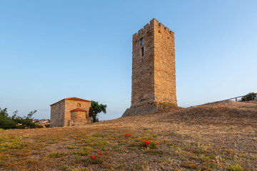 A beautifully preserved ancient Byzantine tower on a hill and a small church nearby at sunrise with bright red poppy flowers in the foreground