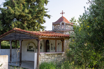 A tiny old orthodox church with stone walls and a tiled roof is surrounded by trees and tall grass