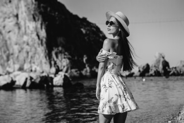 A beautiful young woman in a hat, glasses and a light dress is walking along the ocean shore against the background of huge rocks on a sunny day. Tourism and tourist trips. Black and white.