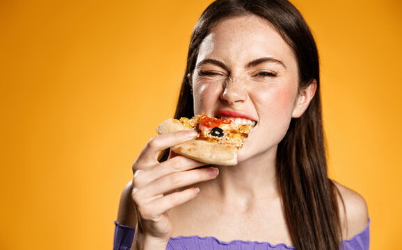 Close up of young woman eating with satisfation, biting pizza slice and looking delighted at camera, try tasty food at pizzeria. Concept of takeaway, fasf-food order or restaurant advertising