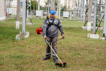 A young man mowing grass on the territory of an electric substation in overalls. Grass cleaning at the enterprise, implementation of fire safety measures