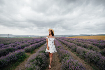 A young beautiful girl in a delicate dress and hat walks through a beautiful field of lavender and enjoys the aroma of flowers. Vacation and beautiful nature.