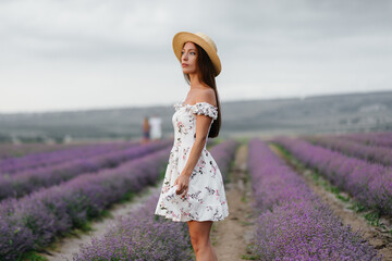 A young beautiful girl in a delicate dress and hat walks through a beautiful field of lavender and enjoys the aroma of flowers. Vacation and beautiful nature.