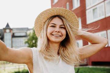 Young smiling cheerful blonde girl in hat making selfie while standing on the street at sunny summer day