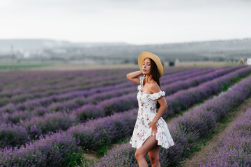 A young beautiful girl in a delicate dress and hat walks through a beautiful field of lavender and enjoys the aroma of flowers. Vacation and beautiful nature.