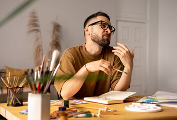 Portrait of male artist sitting at a table coaching in painting classs in studio giving a lesson for students, gesturing. paintbrush in a hand. Lifestyle. Real emotions.