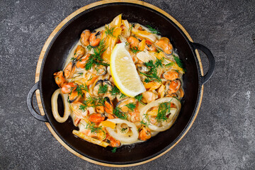 seafood fried in sauce. mussels, shrimp and squid rings with lemon in a hot pan close-up on a dark gray background..