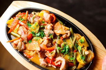 fried potatoes with pork and vegetables in sauce close-up in an oval skillet on a wooden stand