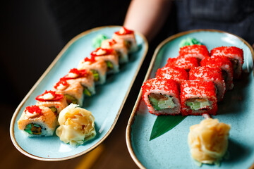 philadelphia roll with baked salmon and cheese, and california roll tuna with red tobiko caviar on blue plates with ginger in the hands of a waiter on a dark background