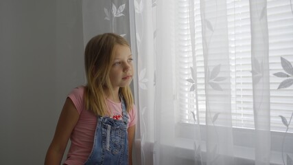 A young school-age girl is at home and looks out the window through a white curtain. Stay at home in quarantine.