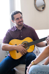 Young and attractive guy playing a romantic song in the guitar to his girlfriend while enjoying a day at home. Woman is surprised and happy listening to his song.