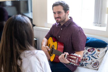Young and attractive guy playing a romantic song in the guitar to his girlfriend while enjoying a day at home. Woman is surprised and happy listening to his song.