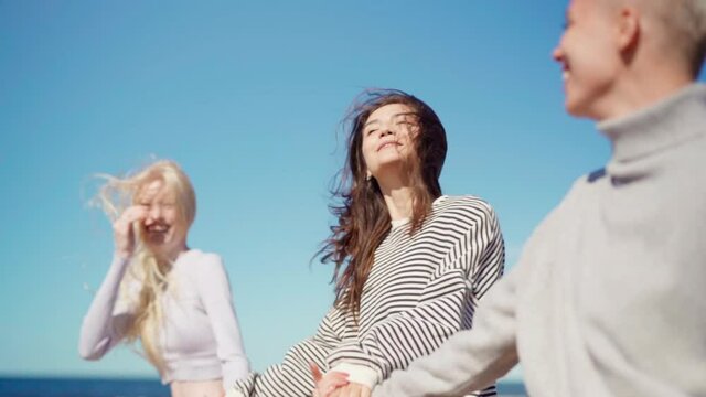 Slow motion shot of three happy young women walking and hopping on beach holding hands against blue sky, their long hair waving in wind. Girlfriends enjoying vacation, freedom and friendship