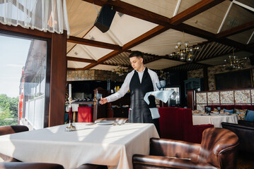 A young male waiter in a stylish uniform is engaged in serving the table in a beautiful gourmet...