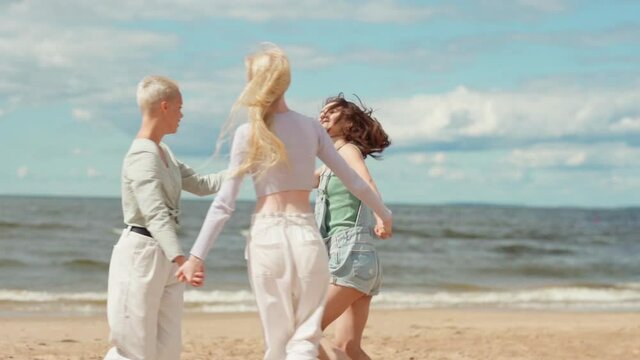 Slow motion panning shot of three happy girlfriends dancing in circle holding hands on beach. Loving women enjoying vacation, freedom and friendship on sea coast