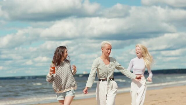 Slow motion shot of two joyful young women walking along sea coast on beach. Happy girlfriend running to join them and taking piggyback ride. Females feeling carefree and happy celebrating life