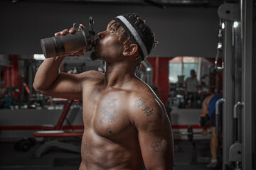 Fototapeta na wymiar African american athletic man with naked torso drinking water or sports nutrition from glass after gym workout