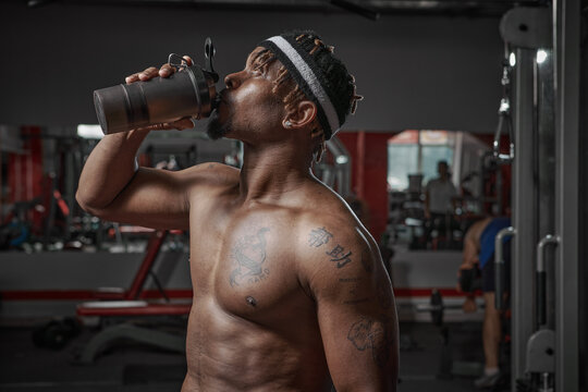 African american athletic man with naked torso drinking water or sports nutrition from glass after gym workout