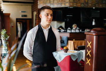 A young waiter in a stylish uniform stands with an exquisite dish on a tray near the table in a beautiful restaurant close-up. Restaurant activity, of the highest level.