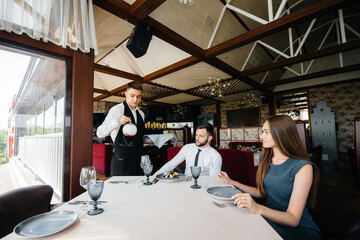 A young waiter in a stylish apron serves a table with a beautiful couple in a refined restaurant....