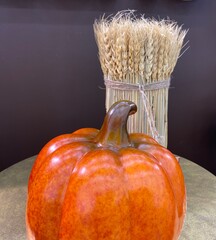 A porcelain orange pumpkin lies on the table in front of a sheaf of wheat stalks on a dark background.