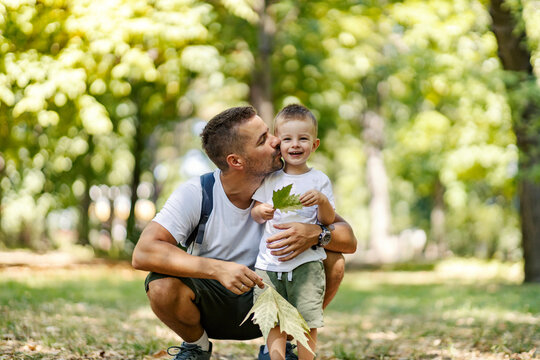 Summer time for family fun in the wood. Father and son dressed in the same clothes fooling around with leaves Dad squats next to the boy and hugs and kiss him. Family moments for remember, hiking