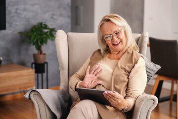 Obraz premium Smiling senior woman sitting in chair at home during lockdown, using tablet and waving. She is having video call with loved ones.