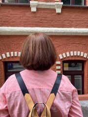 The head of a person (a woman or a man or a teenager) with a short haircut against the background of a red and white brick building in the windows of which cars are reflected.