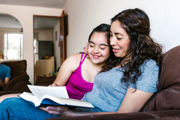 Smiling latin teen girl with down syndrome and her mom reading a book at home, in disability concept in Latin America © Marcos