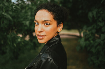 Fashion close up portrait of attractive confident young natural beauty African American woman with afro hair in black leather jacket posing in nature park in green foliage.
