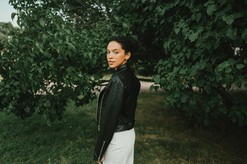 Fashion portrait of attractive confident young natural beauty African American woman with afro hair in black leather jacket posing in nature park in green foliage.