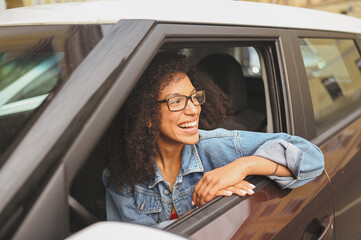 Happy young smiling African American woman black haired driver in glasses sitting in new brown car, smiling looking at camera enjoying journey. Driving courses and life insurance concept.