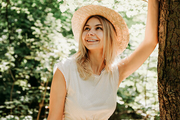 Beautiful girl in a straw hat and stylish clothes stands in a park near a tree. A woman in a white dress near a tree. Close portrait of happy girl