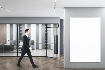 Business man walking in modern concrete glass office interior with empty white mockup poster, daylight, equipment and wooden flooring. Workplace design concept.