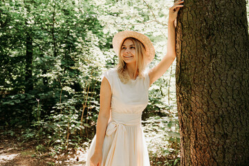 Beautiful girl in a straw hat and stylish clothes stands in a park near a tree. A woman in a white dress near a tree. Close portrait of happy girl