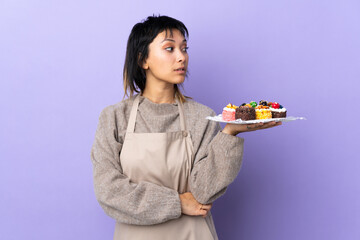 Young Uruguayan woman holding lots of different mini cakes over isolated purple background looking side