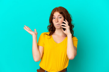 Teenager reddish woman using mobile phone isolated on blue background having doubts while raising hands