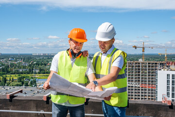 Two smiling mature men in a white helmet stand on the roof, hold a construction plan and inspect the construction site against the background of the city, copying space for text