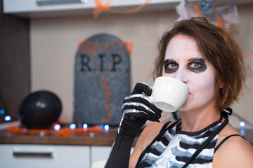 Young contented woman with creepy makeup and in skeleton costume drinks refreshing, relaxing or hot...
