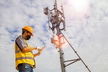 Asian male worker engineer wearing a helmet and safety goggles uses a smartphone to field work near a telecommunication tower controlling cellular electrical installations. - obrazy, fototapety, plakaty