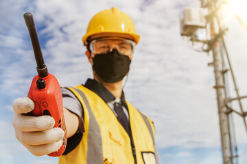 Male worker engineer wearing a helmet and safety goggles uses a walkie-talkie to do fieldwork on a telecommunication tower controlling the installation of cellular electrical equipment.