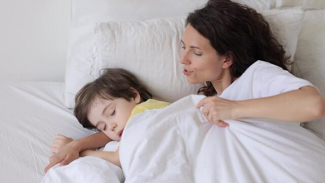 Caring mother cuddling with preschool boy in bed cover sleeping son with warm blanket. Loving young mom comfort little child stroke and caress kid head in bedroom lying together. Tender family morning