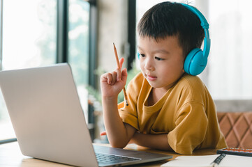 handsome cute Asian boy with looks at the camera while sitting and studying at home on a laptop to study from home during Covid-19 pandemic lockdown. Online education, Homeschooling.