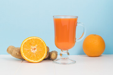 Oranges, ginger and a glass glass with smoothies on a white table on a blue background.