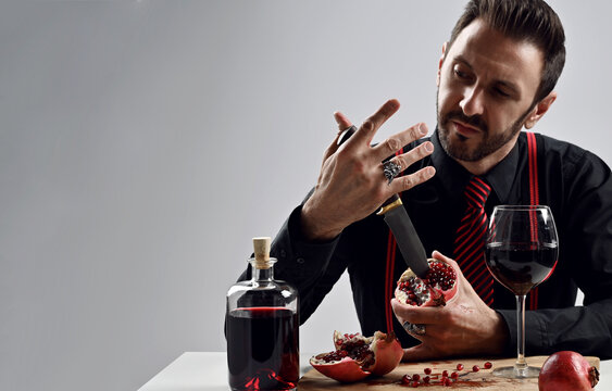 Brutal unshaved man, mafioso in black shirt with striped tie and suspenders sits at table with bottle and glass of juice and sticks a big knife at fresh pomegranate he holds, as if he is a killer