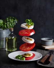 Concept levitation food. Italian caprese salad with tomatoes, mozzarella cheese, basil and pesto sauce fly over the plate on a dark background. Front view